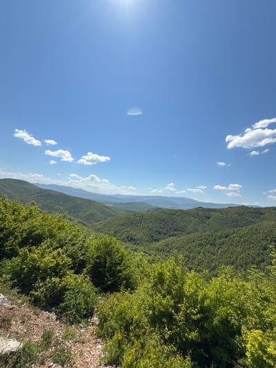 The image shows a landscape of forested mountains under a blue sky with scattered clouds. The foreground features dense green foliage, transitioning into rolling mountain ridges covered in similar vegetation. In the distance, fainter mountains can be seen. The sun is positioned on the left side of the image, causing a bright glare. The location is identified as the city of Lipnitsa, Bulgaria. There are no people or explicit actions visible.