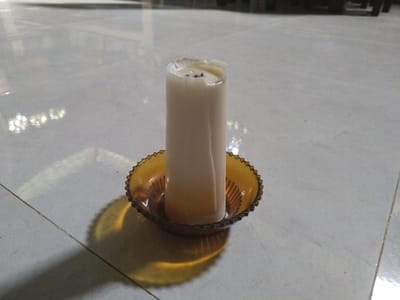 A white cylindrical candle stands upright in an amber-colored glass candle holder, positioned on a light-colored tiled floor. The candle shows clear signs of prior use, with its top edge irregularly melted and a dark wick visible amidst solidified wax. Melted wax has also run down the sides of the candle and collected in the base of the holder.

The candle holder features a decorative design, including a ridged or beaded rim and vertical fluting on its interior base. A prominent shadow of the candle and its holder is cast on the tiled floor to the left, suggesting an artificial light source originating from the right side of the frame. The floor is composed of light grey or off-white tiles, separated by thin, dark grout lines.

The background is softly out of focus, indicating an indoor setting, likely a room, with some faint, shimmering reflections visible on the floor further back. The scene is static, depicting an unlit candle, conveying a quiet, domestic atmosphere. There are no people or visible text present in the image. The location context is Teupinbayu, Indonesia.