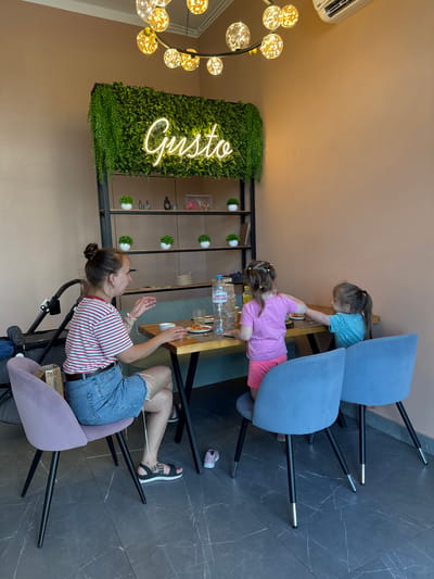 A woman sits at a wooden table in a restaurant with two young girls. The woman, wearing a striped t-shirt and denim shorts, is gesturing with her hands. One girl, in a pink t-shirt and shorts, is reaching across the table. The other girl, in a blue t-shirt, is also at the table. A bottle of water and a slice of pizza are visible on the table. The restaurant has a wall with artificial greenery and a neon sign that reads "Gusto." There are two plush chairs, one lavender and two blue, and a stroller in the background. The floor is dark grey with white marbling. The overall ambiance suggests a casual dining setting in a restaurant located in Patrusheva, Russia.