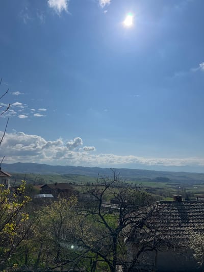 The image presents an elevated daytime view of the rural landscape in Djigurovo, Bulgaria. The upper sky is a clear, bright blue, with scattered white cumulus clouds increasing in density towards the distant horizon. The sun, a brilliant white light source, is positioned in the upper right quadrant, producing a noticeable lens flare. The immediate foreground is framed by a dense array of tree branches, some bare, others exhibiting nascent light green foliage. A prominent deciduous tree with numerous small, dark forms on its bare branches dominates the lower center-right. Partially visible below and to the right is a section of a building featuring a weathered reddish-brown tiled roof. The midground encompasses several rural-style houses with varied roof structures, interspersed within green plots and additional trees. Beyond these dwellings, a landscape of rolling green hills and cultivated fields extends towards the background, terminating in a series of hazy, light blue mountain ranges. No human figures or specific actions are depicted within the scene.