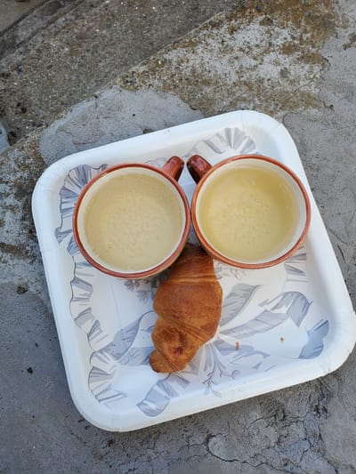 A white plastic tray with a decorative pattern sits on a cracked concrete surface, possibly a stairway. On the tray are two terracotta-colored mugs filled with a light-yellow bubbly liquid. Next to the mugs, a single croissant rests on the tray. The scene appears to be outdoors, given the rough texture of the concrete and the presence of natural light.