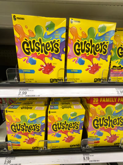 Two shelves of Gushers fruit snacks are displayed in a retail store. The top shelf features two boxes of "Gushers Tropical" and a box of "Gushers Lemonade." Each "Gushers Tropical" box contains 6 pouches and is priced at $2.99. The bottom shelf displays three boxes of "Gushers" fruit snacks, two of which are "Variety Pack Tropical" and one is a "Family Pack" containing 20 pouches. The "Gushers" fruit snacks on the bottom shelf are priced at $9.19. The store environment is indicated by the shelving and product arrangement. The image was captured in Kendall, United States of America.