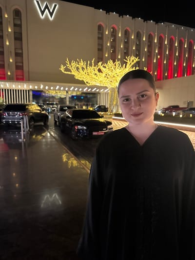 A woman stands in front of the W Hotel at night. The building is lit with red and white lights. A decorative tree with yellow lights stands near the entrance. Cars are parked along the driveway. The pavement reflects the surrounding lights. The woman is wearing a dark garment.