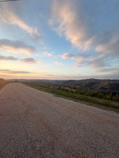 The photograph depicts a rural scene in Castiglione Tinella, Italy, at dusk. An asphalt road stretches into the distance, occupying the right side of the frame and receding towards the center. The left side features a sky filled with clouds, tinted with the soft hues of sunset - light blue, pink, and orange. Along the middle ground, a hillside is visible, covered with vineyards and vegetation, characteristic of the region's winemaking tradition. Small buildings and structures dot the hillside, indicating a settlement. The lighting suggests a peaceful, serene atmosphere as the day transitions to evening. No people or distinct objects are observed.