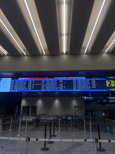 The image shows an airport environment, specifically the Daniel K. Inouye International Airport in Honolulu, United States of America. A flight information display screen prominently features a welcome message in both English and Hawaiian, along with flight schedules, times, destinations, partner airlines, gate assignments, and current status of various flights. In the background, there are airport queuing lanes with retractable belt barriers. Above, there are multiple long LED lighting fixtures. The setting is within the airport terminal, showing a combination of travel-related information and infrastructure.