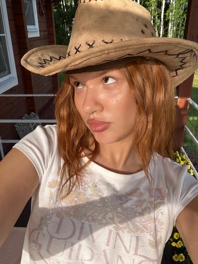 A young woman with red hair and a light tan is wearing a white t-shirt with a floral design and a light brown cowboy hat. She is looking to her left with her mouth slightly open. The woman is standing on a wooden porch with a railing. In the background, there is a red wooden building with windows, trees, and a clear sky. The lighting suggests it is daytime and sunny. The scene appears to be in a rural or suburban area.