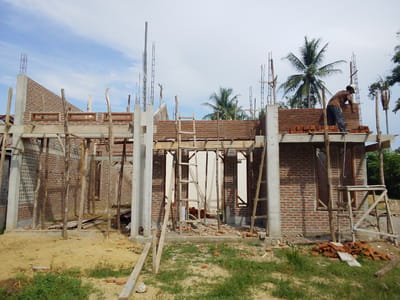 A construction site in Meunasahtambo, Indonesia shows a partially constructed brick building under a cloudy sky. The structure features exposed concrete support pillars and rebar extending upwards. A man wearing a hat and brown clothing stands on a wooden scaffolding, adding bricks to the wall. Several wooden ladders lean against the brick walls. The ground is a mixture of sand, dirt, and scattered brick debris. Some palm trees and other vegetation are visible in the background.