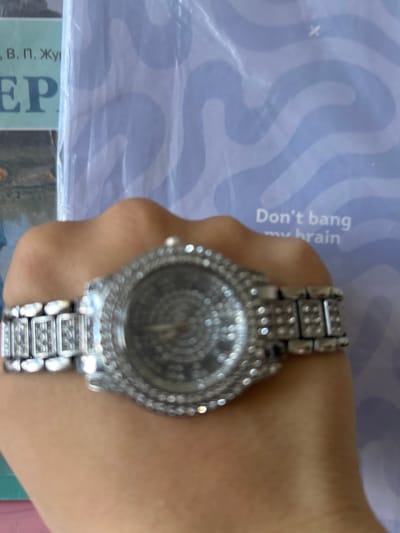 This close-up, vertically oriented image features a person's hand or wrist adorned with a prominent, silver-toned wristwatch. The watch is the main subject, though it is slightly out of focus. It is heavily embellished with numerous clear, sparkling stones, likely rhinestones or simulated diamonds, covering the round bezel and the links of its metal band. The watch face itself exhibits an intricate, possibly textured or patterned design, such as concentric circles.

The setting appears to be indoors, with the hand resting on or near printed materials. The background consists of a pale purplish-blue surface adorned with darker, wavy lines, resembling a patterned cover or sheet. Visible on this background, oriented vertically and partially cut off, is English text that reads, "Don't bang my brain." In the bottom left corner of the image, part of what seems to be a book or magazine cover is visible. This material displays Cyrillic text, including "...В. П. Жу..." and a large Cyrillic letter "Р" (Er), indicating a Russian context. This aligns with the provided location of Dmitrievskaya, Russia. The lighting is ambient, suggesting an indoor environment. No specific activity is depicted other than the wearing of the watch.