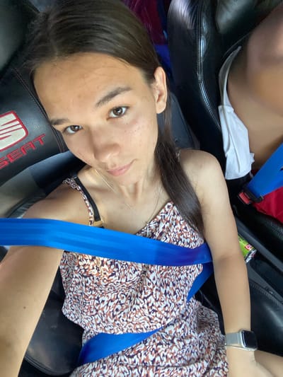 In Berovo, Bulgaria, a young woman with dark hair and brown eyes is seated in a car. She is wearing a floral print dress and a silver smartwatch on her left wrist. A blue seatbelt is fastened across her body. The car seat appears to be black leather with a red "SEAT" logo visible. Another person's shoulder, wearing a white top, is partially visible to her right. A green and white can is also on the seat. The interior lighting suggests it is daytime.