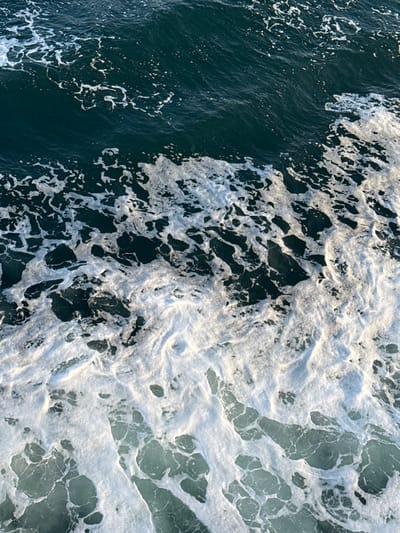 The image displays a close-up, downward view of turbulent water with significant foam, located in Kato Karioutes, Greece. The upper third of the frame shows dark blue-green water with scattered, thin white foam patterns. The middle and lower sections are characterized by dense, irregular patches of frothy white foam, intermingled with lighter blue-green water visible beneath and between the foam clusters. The foam appears brighter, suggesting direct illumination. No individuals or man-made objects are visible within the frame.