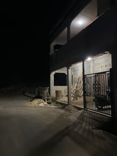 The image depicts a dimly lit scene at night in Landuh, Indonesia. The setting appears to be an outdoor area adjacent to a building under construction or renovation. A metal gate is visible, with wooden planks and debris scattered in front of it. The building itself has exposed brickwork and dark openings that could be windows or doorways. A single light source illuminates a portion of the building's exterior. The ground in the foreground is a paved surface, likely a road or pathway, with some piles of rubble and debris along the edge. The overall mood is somewhat desolate and unfinished, suggesting an ongoing construction project or a neglected space. There is no visible text in the image. The time of day is clearly night, and no specific weather conditions can be inferred. The scene is devoid of any people.