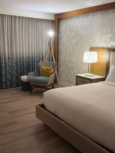 This is a photograph of a hotel room interior, likely in Newport Beach, California. The scene depicts a bedside area and a seating area with a floor lamp. In the foreground, a large bed with neutral-colored bedding is partially visible, with a sliver of the bed frame in tan upholstery. To the right of the bed is a nightstand with a grey drawer and a white lamp with a ribbed shade. Behind the nightstand is a headboard and part of a wall with a textured, geometric pattern in a light neutral tone. To the left of the bed, a rattan armchair with blue cushions and a yellow throw pillow is positioned next to a small, dark cylindrical stool. Behind the chair, sheer curtains with a gradient pattern, transitioning from white at the top to a darker, abstract pattern at the bottom, cover a window. A tripod floor lamp with a light bulb is situated behind the armchair. The flooring is made of wood-look planks. The lighting suggests it is nighttime or the room is dimly lit.