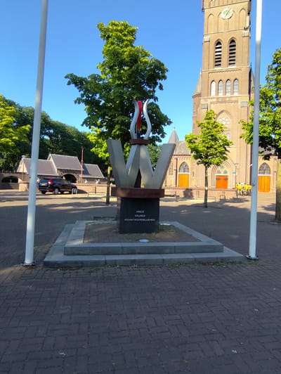 This image depicts a public monument in Schaijk, Netherlands, set against the backdrop of a church and trees. The monument itself is abstract, featuring large, angular metal structures that resemble a stylized "W" topped with a flame-like element in red and silver. A dark granite base supports the sculpture, bearing inscribed text.

In the background, a brick church with a prominent tower and arched windows stands tall. The church has large wooden doors, and several people, some on bicycles, are visible near its entrance, suggesting an ongoing event or activity. To the left of the monument, a modern-looking building with a dark roof is visible, and a black SUV is parked nearby. Lush green trees frame the scene, indicating a pleasant, likely summer day.

The sunlight suggests it is daytime, possibly late morning or early afternoon, casting shadows on the paved ground. The overall mood is serene and somewhat formal, befitting a monument.

The visible text on the monument's base reads:

"DE GEEST VERDRAAGT NIET
VREDE
VRIJHEID
VERANTWOORDELIJKHEID"