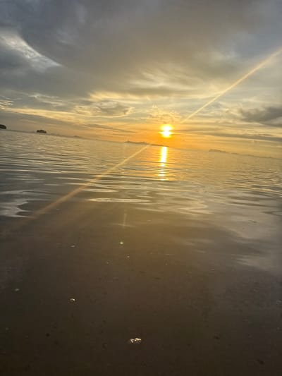 This media file captures a serene sunset over a body of water, likely the sea, given the presence of waves and a distant shoreline. The scene is dominated by the warm, golden hues of the setting sun, which casts a bright, almost blinding glare directly into the camera's lens. This intense light is reflected and refracted by the rippling surface of the water, creating a dazzling effect.
The sky is filled with dramatic clouds, ranging from soft, wispy formations to larger, darker masses that suggest a dynamic weather system. The sunlight pierces through these clouds, creating visible rays and a strong sense of atmospheric depth. A faint, almost iridescent line of light stretches diagonally across the sky and water, possibly a contrail or another atmospheric phenomenon.
On the left side of the frame, a dark, wet surface can be observed, with a few small, reflective circles that might be water droplets or small objects. This area appears to be the shore or a wet, dark ground. In the distance, on the horizon, there are faint outlines of what appear to be islands or landmasses with some vegetation.
The overall mood of the image is peaceful and awe-inspiring, typical of a beautiful sunset. The time of day is clearly evening, as indicated by the low position of the sun and the warm, golden light. The location cues, such as the tropical-looking vegetation on the distant land, along with the provided location context of Baan Pang Ka, Thailand, suggest a coastal setting in Southeast Asia. No visible text is present in the image.
