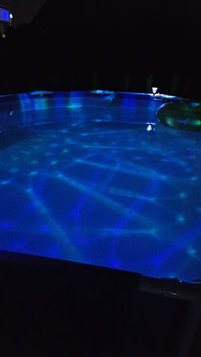 This is a nighttime outdoor photograph of a hot tub. The water is illuminated with blue and green lights, creating a disco ball effect on the surface. A dark green inflatable object, possibly a toy or float, is partially submerged in the water on the right side of the frame. The hot tub appears to be surrounded by dark foliage and structures, suggesting a residential backyard setting. There are no people visible in the image. The location is identified as Ichenhausen, Germany.