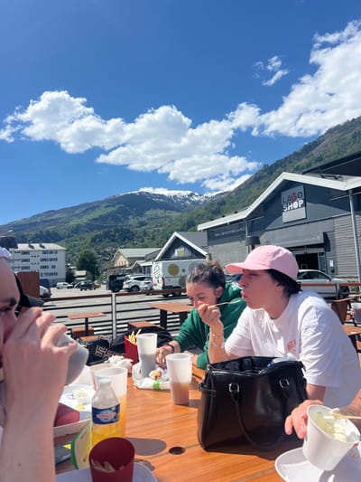 Three people sit at a wooden table on an outdoor patio in Bourg-Saint-Maurice, France. Two women are seated at the table, both wearing sunglasses. The woman to the right is wearing a white shirt and a pink hat and is eating with a fork. The woman to the left is wearing a green hooded shirt. The woman to the right is holding a black purse. There is a man seated to the left of the image who is not fully visible. The table is covered in various eating items and utensils, including empty containers of takeout food, utensils, cups, and a bottle of orange soda. In the background, buildings and a mountain are visible. There is a blue sky with white clouds above.