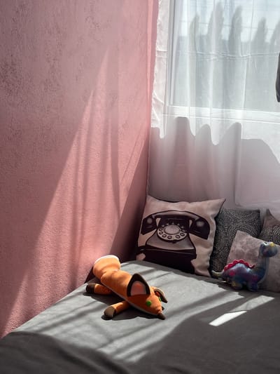 The image displays an indoor scene, likely a bedroom or a cozy living area, illuminated by natural sunlight. The perspective is from above, looking down into a corner of the room.
On the left side of the frame, occupying the upper portion, is a window covered by sheer, white, translucent curtains or blinds. The fabric appears somewhat ruffled or folded, allowing diffuse light to enter.
The bottom left portion of the image is dominated by a textured, matte pink surface, which could be a wall meeting a floor, or a large rug. This surface has an uneven, slightly rough finish.
On the right side of the image, a grey upholstered surface is visible, suggesting a bed or a sofa. Several items are arranged on this grey surface:
- A white decorative pillow with a prominent black illustration of a vintage rotary telephone, including the handset and a numbered dial.
- Behind this pillow, other pillows with intricate grey and black patterned fabric can be seen.
- Near the top of the grey surface, resting among the pillows, is a small, colorful figurine, possibly a dinosaur or dragon, with iridescent blue, purple, and pink scales.
- Further down on the grey surface, an orange and brown stuffed animal, resembling a fox or a deer with small green eyes, lies with its head pointed towards the lower left.
The room is bathed in strong, natural sunlight streaming through the window. This light casts distinct, elongated shadows across both the pink surface and the grey upholstery. The shadows from the window coverings create a striking wavy pattern on the pink surface, indicating the sun's low angle, likely suggesting either morning or late afternoon.
The overall impression is one of warmth, tranquility, and domestic comfort. There is no discernible activity taking place, and no text is visible in the image.