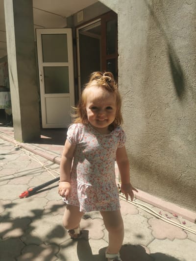 A young child with blonde hair styled in two small buns is standing on a paved outdoor area. The child is wearing a floral print dress and sandals and is smiling at the camera. To the left of the child is a column and a partially open door with glass panels. The background consists of a textured concrete wall and the ground is paved with interlocking tiles. A shadow is cast on the wall from the right side of the frame. The scene appears to be a residential area in Carbuna, Moldova, possibly a courtyard or porch.