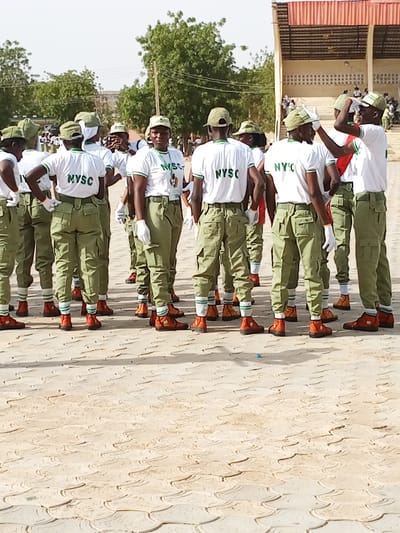 The image shows a group of young adults, likely corps members participating in the National Youth Service Corps (NYSC) program in Ajiwa, Nigeria. They are dressed in the standard NYSC uniform of olive green trousers and white t-shirts with green trim. Most of them are also wearing light green caps and white gloves. They are standing in formation on a paved outdoor area with a distinctive patterned surface. In the background, there is a covered seating structure with a red roof, and trees suggesting a warm climate. The lighting indicates it is daytime, likely under a bright sun, given the strong shadows cast on the ground. The individuals appear to be attentive and organized, suggesting a parade, assembly, or training session. The overall mood seems formal and disciplined, fitting for a structured event. The visible text on their shirts reads "NYSC".