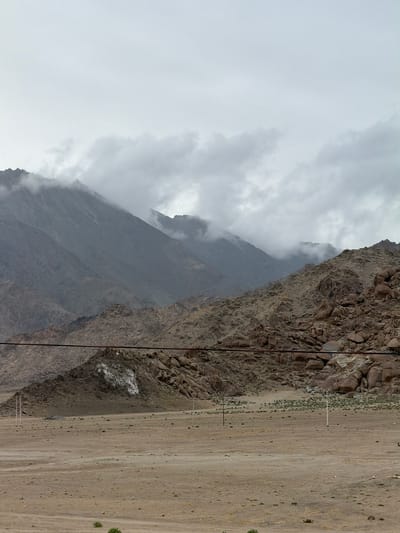 A horizontal, medium shot captures a barren, dusty landscape at the base of imposing, rocky mountains under a cloudy sky in Chumkar, India. The foreground is a vast expanse of tan-colored dirt, sparsely dotted with scrubby green vegetation. Several utility poles, thin and white, stand scattered across the plains, with dark electrical wires stretched taut between some of them. The middle ground is a steep incline of rough, brown rocks and boulders, some appearing quite large. A prominent feature in this section is a large, irregular white patch on a dark rock face. In the background, the mountains rise dramatically, their peaks partially obscured by swirling grey clouds that hug their slopes. The overall impression is one of a remote, arid, and somewhat desolate environment.