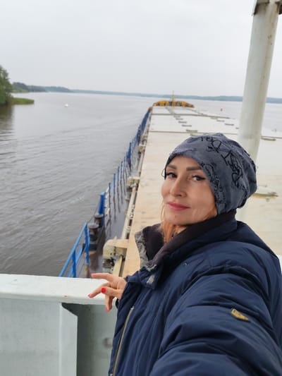 A woman with orange hair, wearing a navy blue puffer jacket and a grey beanie, stands on a ferry in Goritsy, Russia. She is looking towards the camera with a slight smile, her head tilted. Her right hand is raised, with bright red nail polish visible. The ferry is on a calm body of water, with a distant shoreline and a hazy sky. The background shows the length of the ferry, with yellow structures at the far end.