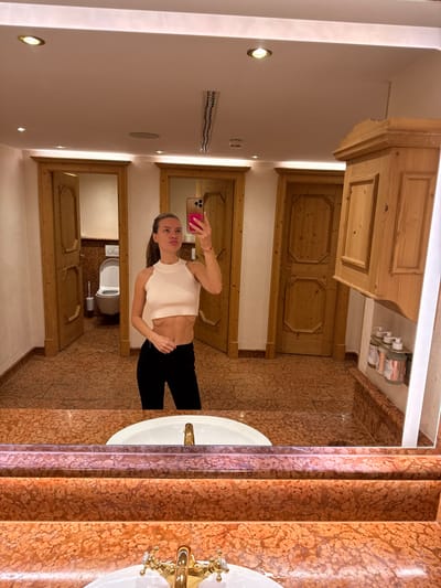 The image captures a mirror selfie of a young adult female inside a restroom located in Going am Wilden Kaiser, Austria. The female, wearing a white sleeveless crop top that exposes her midriff and dark pants, holds a pink smartphone in her right hand, pointed at the mirror to capture the photograph. She stands before a reddish-brown marble vanity featuring an integrated white sink and a decorative gold-toned faucet.
Reflected in the mirror, behind the subject, are three light-colored wooden doors with carved panels. The leftmost door is partially ajar, revealing a white wall-mounted toilet and a toilet brush holder within an individual stall. To the right of the reflection, a matching wooden wall-mounted cabinet with multiple panels is visible above a shelf containing several bottles of toiletries. The restroom floor is tiled with reddish-brown, patterned tiles, and the white ceiling features multiple recessed circular spotlights providing bright illumination, along with a linear air vent.