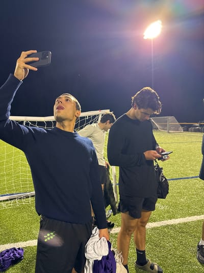 The image depicts three males on an outdoor artificial turf sports field at night, illuminated by stadium floodlights, in Newton, United States. The exact facility within Newton is not discernible. In the foreground left, a male in a navy blue long-sleeved sweater and black athletic shorts holds a dark-cased smartphone aloft with his right hand, pointing it towards himself and slightly upward, as if taking a selfie or video. His tongue is partially extended. He holds a bundle of white and purple fabric in his left hand. To his right, another male in a black long-sleeved shirt, black athletic shorts, dark socks, and light-colored slide sandals stands looking down at a smartphone held in both hands. A black bag hangs from his left shoulder. In the background, partially obscured by the foreground figures, a third male in a light-colored long-sleeved shirt and dark shorts stands near a white-framed soccer goal net. A bright stadium floodlight is visible in the upper right quadrant of the image. The field features white demarcation lines.