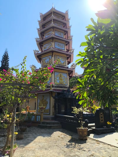 The image captures a tiered pagoda structure situated in Tay Nha Trang Ward, Vietnam, on a bright, sunny day. The pagoda, adorned with intricate golden reliefs depicting figures and scenes, rises prominently against a clear blue sky. Lush green foliage and vibrant pink bougainvillea flowers surround the base of the pagoda, partially obscuring the view of the lower levels and surrounding structures. Several potted plants and dark, ornate statues or altars are visible in the foreground and mid-ground, contributing to the temple's ambiance. The ground is paved with stone or concrete, with patches of dry grass and shadows cast by the trees. The overall scene suggests a serene temple courtyard under a warm, sunlit sky.