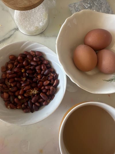 Here is a concise description of the image:
The image shows a tabletop with several food items. There's a bowl containing three brown eggs, a bowl filled with peanuts, and a mug filled with a light brown liquid, possibly coffee with milk. A jar with a wooden lid, potentially containing sugar, is also visible. The scene appears to be a simple breakfast or snack arrangement. Location information (Черёмушки, Russia) is not evident in the image itself.