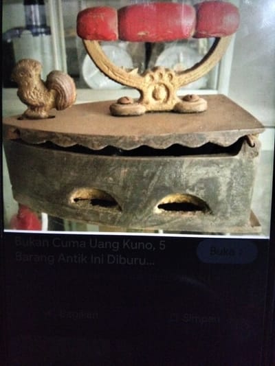 This is a close-up, eye-level, outdoor shot of an antique charcoal iron displayed on a weathered wooden surface. The iron is made of dark metal and has a decorative rooster figure perched on its lid, which is embellished with red ceramic handles. The iron has a wavy edge and two oval-shaped openings on its side. The background is blurred and suggests a display area, possibly a market stall or antique shop, with glimpses of other items and a reflective surface. A portion of a digital screen with Indonesian text and icons is visible at the bottom, indicating the image might be a screenshot or viewed on a device. There is no visible presence of people, and the environment appears to be a commercial or exhibition setting, not specifically within Kualaraja, Indonesia.