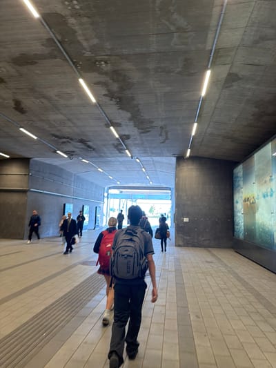 The image depicts a modern pedestrian passage or tunnel, likely part of a public transportation facility or large building in Brossard, Canada. The perspective looks down the length of the passage towards a bright opening at the far end.
Multiple people are walking away from the camera, moving towards this bright exit. In the foreground, a person wearing dark pants and a grey shirt, carrying a grey backpack, is central. The brand "SCOOTS" is clearly visible on this backpack. Behind them, another individual wears a red jacket and dark pants. Further down the passage, several other people are seen, all generally moving in the same direction.
The left wall is made of dark, textured concrete, adorned with multiple horizontal linear LED or fluorescent lights that illuminate the space. The right wall and the floor are constructed from lighter-colored, rectangular paving stones or tiles. Darker longitudinal stripes are integrated into the floor, possibly for visual or tactile guidance. The ceiling is high and dark. Towards the far end of the passage, a large, rectangular panel is mounted high on the wall or ceiling, displaying what appears to be an abstract artwork or reflective image, perhaps depicting water or a landscape. The architecture is stark, modern, and functional. No specific time of day or weather is evident from within the enclosed passage.
