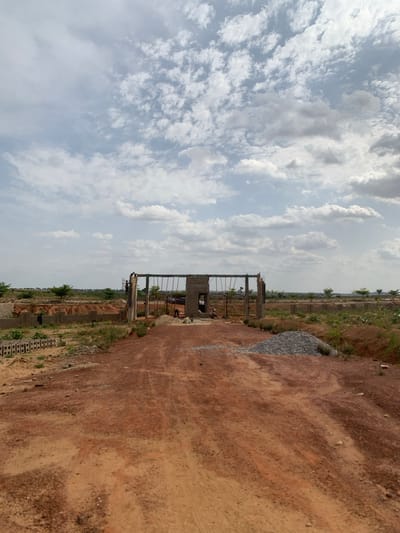 A photograph captures a construction site in Kujama, Nigeria. The sky, filled with white, layered clouds, occupies approximately half the frame. Below, an expanse of reddish-brown soil dominates, with a partially constructed metal gate structure positioned centrally. The gate is not complete, and construction is ongoing. Scattered grey gravel lies to the right of the gate, amidst the reddish-brown earth. In the distance, vegetation and other structures, possibly dwellings, are visible, suggesting a developing urban or peri-urban landscape. No people are immediately visible within the frame.