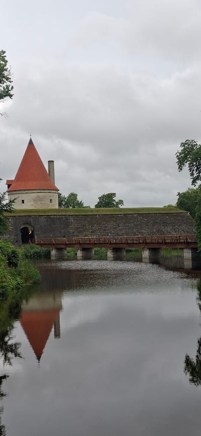 The image shows the Kuressaare Castle in Kuressaare, Estonia. The castle features a distinctive conical red roof and a stone tower. A wooden bridge spans a moat in front of the castle, leading to an arched entrance. The moat reflects the castle and the overcast sky. Green trees and foliage surround the castle, and the water's surface is rippled, distorting the reflections. Two individuals are visible on the bridge. The overall scene depicts a historic fortress under a cloudy sky.