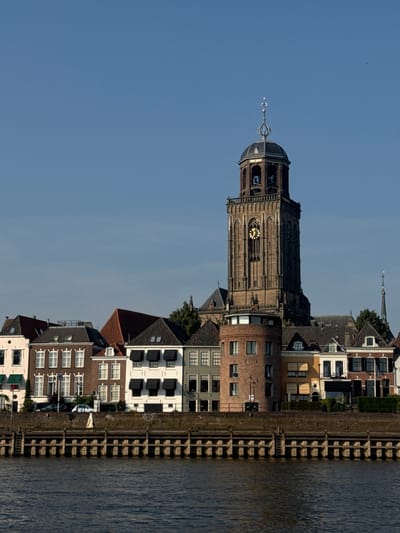 The image shows a wide shot of the IJssel river with a city skyline on the opposite bank in Deventer, Netherlands. In the foreground, the dark, rippling water of the river takes up the bottom third of the image. The middle ground features a stone embankment with wooden pilings, supporting a row of historic buildings. Dominating the skyline is the towering Great Church of Saint Lebuinus (Grote of Lebuïnuskerk), a prominent Gothic cathedral with a large, square bell tower featuring a clock face. Adjacent to the church are various other buildings with traditional Dutch architecture, characterized by steep gabled roofs and varied colored facades. The sky above is a clear, bright blue. There are no people visible in the image.