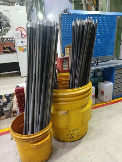 In an industrial setting, possibly a workshop or warehouse in Қопа ауылдық округі, Kazakhstan, a large number of long, threaded metal rods are stored upright in two yellow containers. The rods are dark gray and appear to be of uniform size and length. The container in the foreground is a wide, short yellow drum, while the container behind it is a taller, more cylindrical yellow bin.
In the background, a blue pegboard with a metal frame is mounted on a wall, holding various tools or components. To the left of the pegboard, a white industrial machine is partially visible, with some signage and cables attached. There are also several white and red plastic containers and wooden pallets scattered around the floor. The floor itself is concrete, with yellow and red lines marking pathways or boundaries. The lighting suggests an indoor environment with overhead fluorescent lights. No people are visible in the image.
