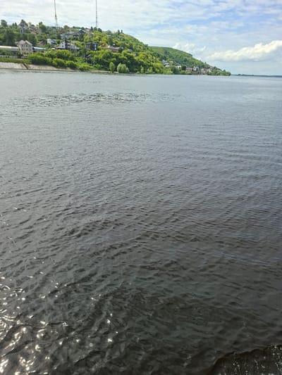 The image captures a wide expanse of a dark, rippling body of water, likely a river, under a partly cloudy sky. In the background, a verdant, hilly landscape is visible, dotted with numerous houses and buildings that form a settlement along the shoreline. The buildings are varied in size and style, with red, brown, and white roofs prominent amongst the greenery. Two tall radio towers are also visible on the highest point of the hill. The shoreline itself appears to be reinforced in some sections with concrete structures. The overall impression is of a tranquil, residential area situated along a significant waterway. The location is identified as Verkhny Uslon, Russia.