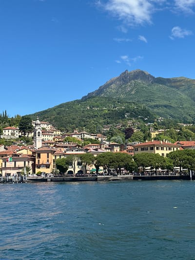 This panoramic image captures a scenic view of Loveno, Italy, nestled against a steep, verdant mountain range alongside a wide body of blue water, likely a lake. The scene is depicted on a bright, clear day, under a blue sky dotted with scattered white clouds.
On the right side of the frame, the calm, deep blue water stretches out, exhibiting subtle ripples across its surface. Along the water's edge, a lively promenade or waterfront area is visible, lined with lush green trees and what appear to be numerous outdoor restaurant or cafe setups. Small, indistinct figures of people can be seen enjoying the waterside setting, suggesting leisure activity.
The town of Loveno occupies the left and central parts of the image, featuring a cluster of multi-story buildings. These structures display characteristic warm-toned facades, including shades of yellow and orange, topped with traditional terracotta roofs. A distinctive white church with a prominent dome-shaped bell tower stands out amongst the other buildings. The town extends upwards along the lower slopes of the mountain, which is heavily forested with various trees, revealing rugged, rocky terrain towards its summit. An Italian flag is visible, proudly displayed on one of the buildings along the waterfront. The overall impression is one of a tranquil, sunny day in a picturesque Italian lakeside town.