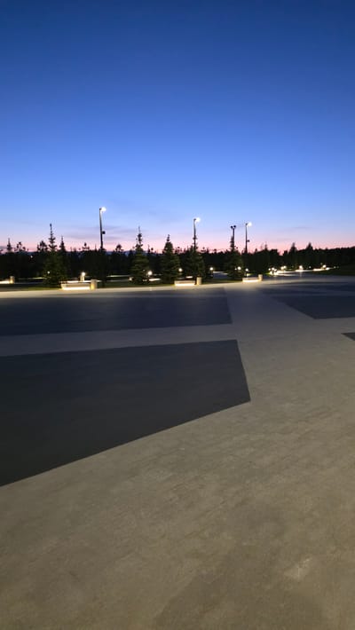 The image shows an outdoor scene at dusk, possibly in the Izhora area of Verevskoe S/P, Russia. The right side of the image is dominated by a concrete or paved surface with darker gray square or rectangular patterns. Along the horizon line, there are trees illuminated by streetlights. The sky transitions from a deep blue above to a lighter pinkish-purple near the horizon. There are no visible people or discernible interactions taking place. The image appears to be taken at ground level.