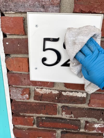 A close-up, eye-level shot captures a gloved hand wiping a white house number sign attached to a red brick wall. The hand, encased in a bright blue latex or nitrile glove, holds a light-colored, possibly white or off-white, terry cloth towel. The towel is pressed against the upper-middle section of the white rectangular sign, obscuring the top portion of the black house numbers.
Visible on the sign are the distinct black numbers "1" and the top and side curve of a "5," suggesting the full number might be "15" or a similar combination starting with "15". The numbers appear to be raised or applied onto the white surface. Small, silver-headed screws are visible in the corners of the white sign, securing it to the brickwork.
The background consists of a textured red brick wall with gray mortar lines. The bricks show varying shades of red and brown, with some exhibiting white efflorescence or weathered patches. At the very bottom of the frame, a segment of a light blue painted surface or trim is visible, providing a contrasting color element. The lighting is even, suggesting an outdoor setting during daytime, possibly under cloudy conditions or in shade. The image conveys a sense of routine maintenance or preparation for a delivery/service at a residence or building in Hampton, United States of America.