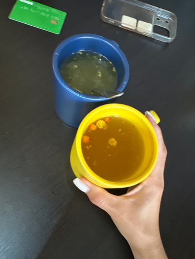The image depicts a dark, textured tabletop with several objects arranged on it. In the foreground, a person's hand with neatly manicured white nails is holding a bright yellow mug filled with a golden-colored liquid containing small, orange and yellow particles. To the left and slightly behind the yellow mug, a blue mug is visible, also containing a similar liquid with scattered particles. A silver spoon is partially submerged in the blue mug. In the upper left corner, a green card with white text and a small brown chip is partially visible. Further back, a clear plastic phone case with two square, light-colored objects inside is present. The scene is illuminated by overhead lighting, casting soft shadows. The overall setting suggests a casual indoor environment, possibly a home or office.