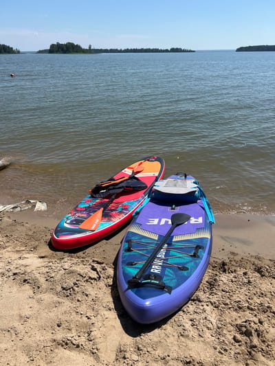 Two paddleboards, one red and orange with a colorful graphic of a space scene and a person, and another purple and blue with a geometric pattern and the word "RAVE" prominently displayed, are resting on a sandy beach next to a body of calm water. The red paddleboard has a life vest and a paddle placed on top of it. The purple paddleboard has a paddle resting on it and a small white platform attached to its center. In the distance, a large body of water stretches out towards a tree-lined shore under a clear blue sky. The scene is consistent with a recreational area such as Borovoye, Russia, known for its natural beauty and water activities. A person is visible in the water in the far left of the frame.