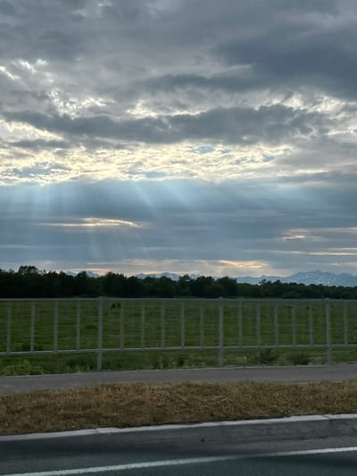 The image depicts a landscape scene. The sky is filled with dramatic clouds, with rays of sunlight piercing through them. In the foreground, a grassy field is visible behind a metal fence. Beyond the field, a line of trees forms a dense border, and in the distance, mountains can be seen. The overall impression is one of a natural, open environment under a dynamic sky. There are no people or specific urban elements of Кеткино, Russia identifiable in the image.