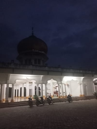 The image depicts a mosque at night or late dusk/early dawn, located in Pulo Lhok, Indonesia. The dominant feature is the mosque, with its large, dark, bulbous dome and a distinct finial silhouetted against a dark blue-black sky. The upper section of the mosque, beneath the dome, is also dark and blends into the night.

In stark contrast, the lower level of the mosque is brightly illuminated by artificial lights, highlighting its white pillars and decorative arches. This section features a wide, elevated platform accessible by steps. Inside the brightly lit area, several people are visible, seated on the floor, likely engaged in prayer or religious contemplation.

In the foreground, a paved courtyard or parking area is visible, where at least five motorbikes are parked. The motorbikes are dark and mostly in silhouette, but their presence suggests attendees have arrived for worship. The sky is dark and mostly clear, with subtle variations in tone, indicating a late hour. No specific weather conditions are discernible beyond the darkness. The architectural style, with its dome, arches, and pillars, is characteristic of Islamic religious buildings. There appears to be some dark, indiscernible text or design within one of the arched sections above the pillars inside the mosque. The overall atmosphere is one of tranquility and devotion.