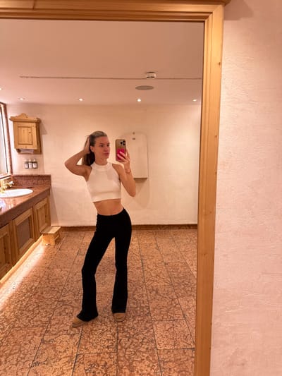 A young adult female is depicted in a full-body mirror selfie taken in what appears to be a private or public restroom or changing room within an establishment in Going am Wilden Kaiser, Austria. The female, with blonde hair pulled back in a ponytail, wears a white ribbed sleeveless crop top, black flared trousers, and tan Ugg-style boots. She holds a pink-cased smartphone in her right hand, angled to capture her reflection, while her left hand is positioned behind her head, fingers touching her hair.
The reflected environment features light pink-toned, textured walls and a floor tiled with reddish-brown and tan patterns. To the left, a light wooden vanity unit with a reddish-brown marble countertop supports a white oval sink and a silver faucet. Above the sink, a small wooden wall cabinet and three white wall-mounted dispensers are visible. A small wooden step stool rests on the floor next to the vanity. The ceiling contains recessed lighting and a smoke detector or sprinkler head. To the right of the reflection, an arched alcove or structural feature is present on the wall. The room is uniformly illuminated by artificial light.