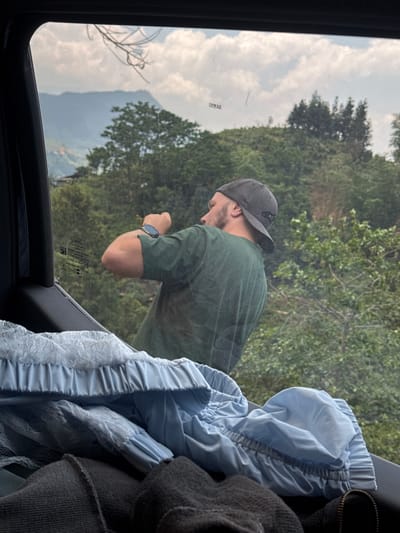 The scene is viewed from inside a vehicle, likely a car, looking out the window. A man with a beard and a grey baseball cap, wearing a green t-shirt, is reflected in the window, appearing to look out at the scenery. He has a watch on his left wrist. The environment outside the window is a lush, mountainous area, likely in a rural setting. The reflection suggests the presence of trees and possible hills or mountains, with clouds in the sky. The lettering printed on the car window is visible. In the interior of the car, there are folded light blue clothes with lace trim and a dark gray, fleece-like garment, partially obscuring the view. Based on the text from the car window, this image was taken in Hoàng Su Phì Commune, Vietnam.