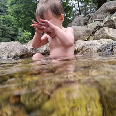 A baby sits in shallow, clear water near the bank of a stream. The baby's hands are held up to their face, covering their eyes, and water drips from their fingers. The baby is partially submerged, with their torso and legs visible above the water. The surrounding environment consists of large, moss-covered rocks and lush green trees. The water is shallow enough to see the rocky bottom with some greenish algae.