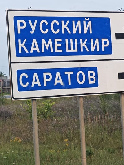 A road sign with white text on a blue background indicates directions to two locations. The top line reads "РУССКИЙ КАМЕШКИР" and the bottom line reads "САРАТОВ". The sign is supported by two metal poles. The background consists of tall grass and some trees, suggesting a rural or roadside setting. The lighting indicates it is daytime.