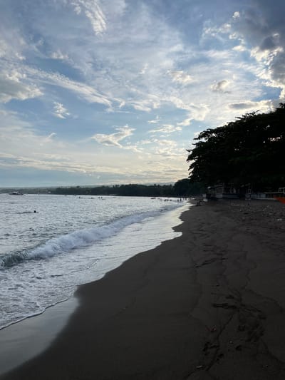 The image depicts a beach scene in Dauin, Philippines, under a sky filled with scattered clouds. The dark sand of the beach occupies the foreground, leading to the ocean. Waves gently break on the shore, with foam spreading across the wet sand. In the water, several people are visible, some swimming and others engaged in water activities. A boat is also present in the distance. Along the edge of the beach, a line of trees provides shade, and structures that appear to be huts or small buildings are situated beneath them. The overall atmosphere is serene, suggesting a leisurely day at the beach.