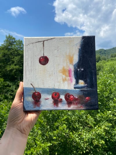 A square canvas painting of cherries is held in a person's hand against a backdrop of lush green trees and a blue sky with clouds. The painting depicts several red cherries on a white and blue surface, with one cherry suspended from a stem. The background of the painting is abstract, with dark blue and yellow hues. The painting is rendered in an impressionistic style with visible brushstrokes. The scene is set outdoors, in Lipniki, Russia, as indicated by the natural environment.