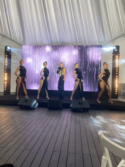 A group of five women are performing on a stage in what appears to be an outdoor event, possibly a wedding reception or corporate event. The stage is set up on a wooden floor, and behind the performers is a large LED screen displaying a shimmering, starry background. There are spotlights and stage lighting illuminating the scene.
The performers are dressed in similar, glamorous outfits. Four of the women are wearing black, sequined dresses with high slits, and the woman in the center is wearing a black jumpsuit with a sequined bodice and black sleeves. She is also holding a microphone and appears to be singing. The other four women are dancers, striking poses and performing synchronized movements.
The event is taking place under a large white tent, suggesting it is an outdoor venue. The overall atmosphere is festive and professional, indicating a high-quality production. The image was taken at Калодзішчанскі сельскі Савет, Belarus.